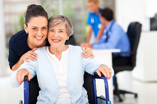 CNA in blue scrubs standing behind elderly woman smiling in wheelchair